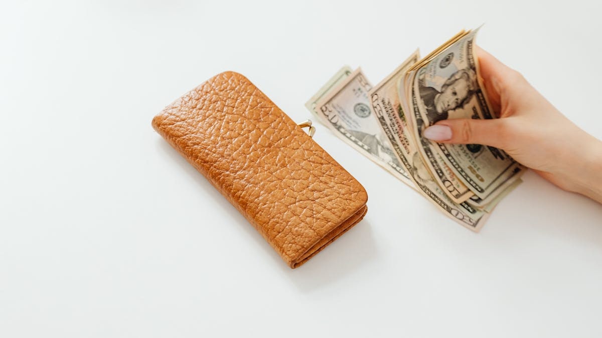 A hand counting American dollar bills next to a brown leather wallet on a white surface.